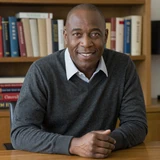 David, a smiling man in charcoal sweater seated at a wooden study desk lit by warm lamplight